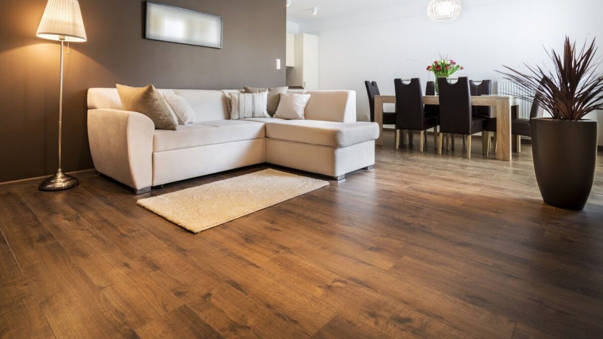A modern living room featuring elegant wooden flooring, a beige sectional sofa, a stylish floor lamp, and a dining area with a wooden table and black chairs. A decorative plant adds a touch of greenery to the space.
