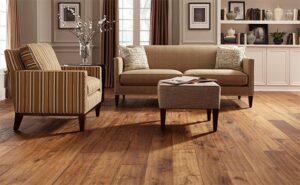 A cozy living room with warm-toned hardwood flooring, striped armchairs, and a comfortable sofa, complemented by soft, natural light streaming through large windows.