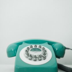 A vintage turquoise rotary dial telephone placed on a white surface against a light gray background.