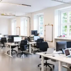 Modern open-plan office with rows of desks, computers, and ergonomic chairs in a bright, spacious room with large windows and natural light