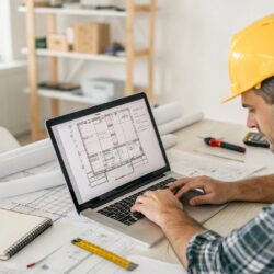 Construction professional wearing a yellow hard hat working on a laptop with building plans and blueprints displayed on the screen, surrounded by drafting tools and documents on a desk.